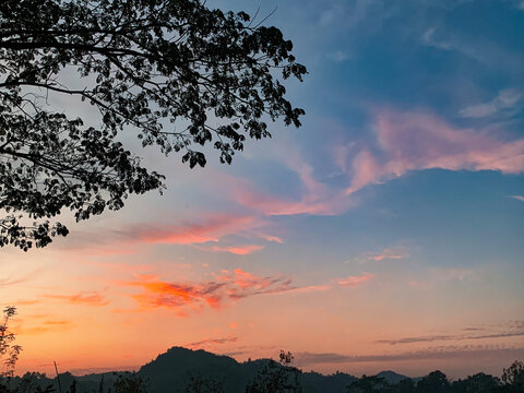 View of vibrant sunset sky with orange and pink clouds over the hilly landscape of Nhila in Teknaf, framed by silhouetted tree branches in Cox's Bazar, Chittagong Division, Bangladesh.