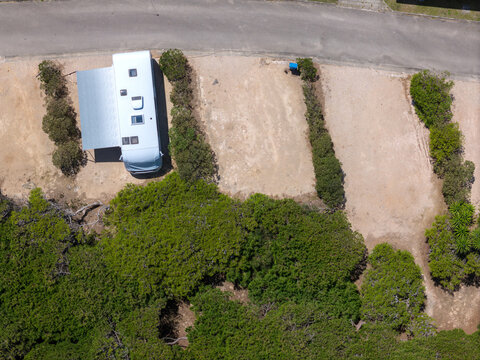 Aerial view of a white camper van with an extended awning parked in a gravel lot surrounded by green bushes and trees, Cascais, Lisbon, Portugal.