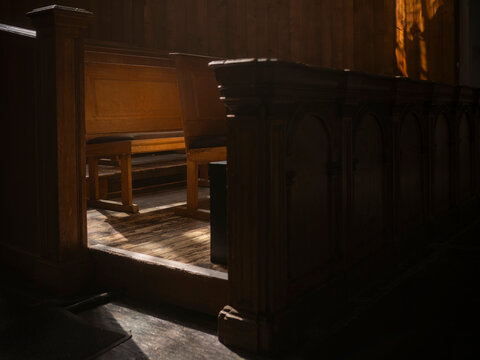 Wooden pews and a panelled enclosure caught in a shaft of light with a weathered plank floor, moody historic church interior atmosphere.