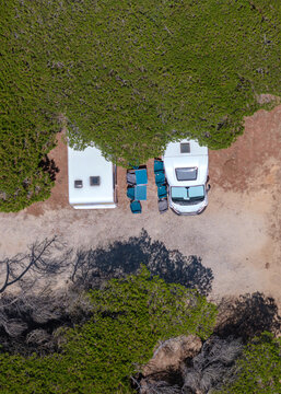 Aerial view of a white camper van parked on a gravel lot with camping chairs and a table set up under the shade of nearby trees in Cascais, Lisbon, Portugal.