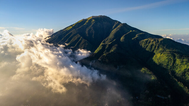 Aerial view of Mount Merbabu with lush green slopes partially covered by dramatic clouds under golden sunlight in Selo, Central Java, Indonesia.