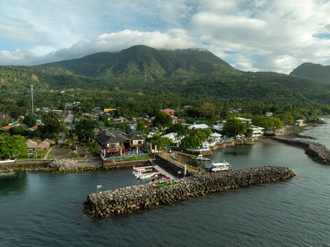 Aerial view of the coastal town of Mambajao with its rocky breakwater, small boats moored at the jetty, and lush green mountains in Mambajao, Camiguin, Philippines.