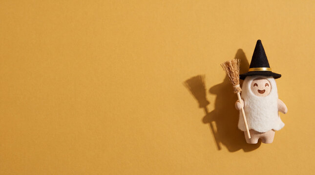 A cute little felt ghost wearing a black witch hat and holding a broom on a gold-toned background with copy space.