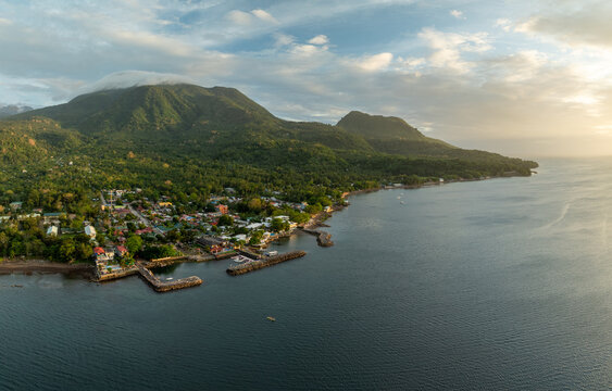 Aerial view of the coastal town of Mambajao nestled between lush green mountains and the calm sea, featuring small piers and residential rooftops in Mambajao, Camiguin, Philippines.