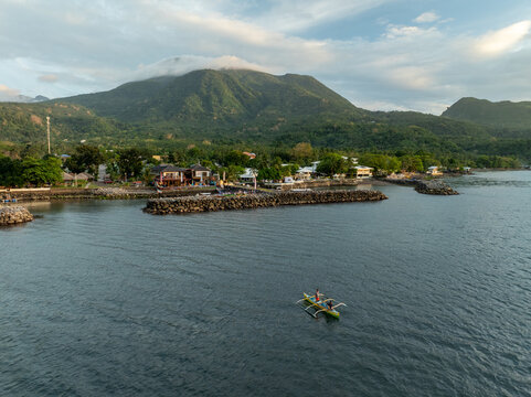 Aerial view of a traditional outrigger boat sailing on the calm coastal waters near the lush green mountains and shoreline of Mambajao, Camiguin, Philippines.