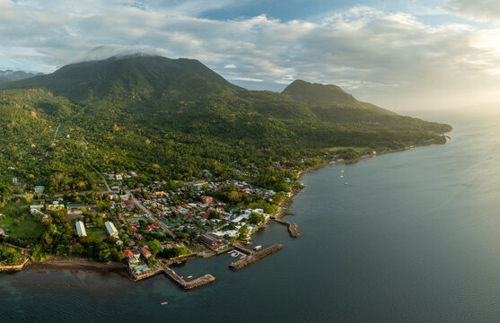 Aerial view of the coastal town of Mambajao with lush green mountains, dense tropical vegetation, and a small harbor along the coastline in Mambajao, Camiguin, Philippines.