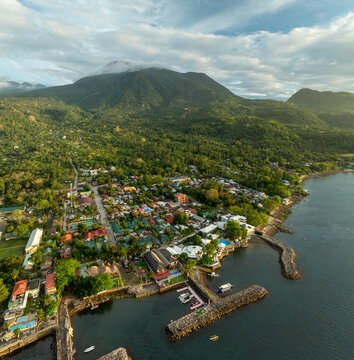 Aerial view of the coastal town featuring a stone pier, colorful rooftops, lush tropical vegetation, and mountains under a cloudy sky in Mambajao, Camiguin, Philippines.