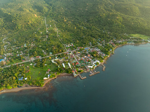 Aerial view of the coastal town of Mambajao with lush green hills, palm trees, and small piers extending into the sea in Mambajao, Camiguin, Philippines.