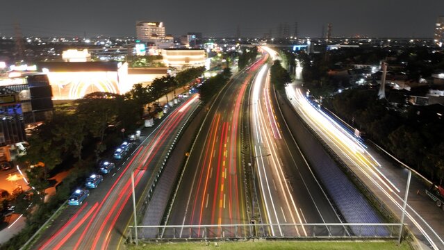 Aerial view of Jakasampurna highway at night with long exposure light trails from vehicles, illuminated urban buildings, and city lights in Bekasi Barat, West Java, Indonesia.