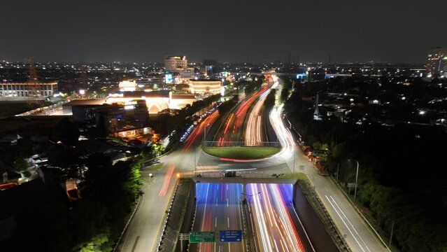 Aerial view of the Jakasampurna highway interchange at night with vibrant light trails from passing vehicles, surrounded by the illuminated urban landscape of Bekasi Barat, West Java, Indonesia.