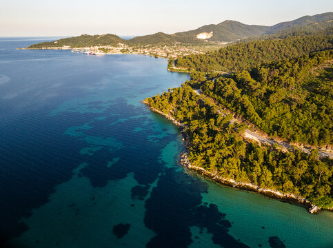 Aerial view of the lush green coastline and turquoise waters of Thasos island with a winding coastal road surrounded by dense pine forests in Thasos, Greece.