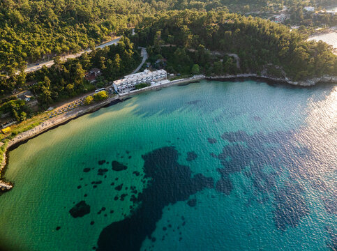 Aerial view of the coastline featuring turquoise waters, a sandy beach, a resort hotel, and surrounding pine forests under warm golden hour light, Thasos, Greece.