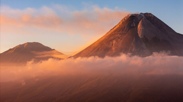 Aerial view of Merapi volcano at sunset, with golden light illuminating the volcanic peak and clouds drifting around the slopes in Pakem, Yogyakarta, Indonesia.