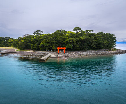 Aerial view of the iconic red torii gate at Mihama Beach, surrounded by lush green trees and rocky coastline along the clear blue waters of Suruga Bay in Numazu, Shizuoka, Japan.