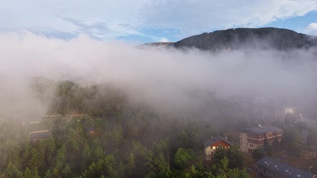 Aerial view above thick fog rolling through a mountain valley near La Molina, Catalan Pyrenees, Spain, with village buildings emerging below cloud line and ski slope visible above