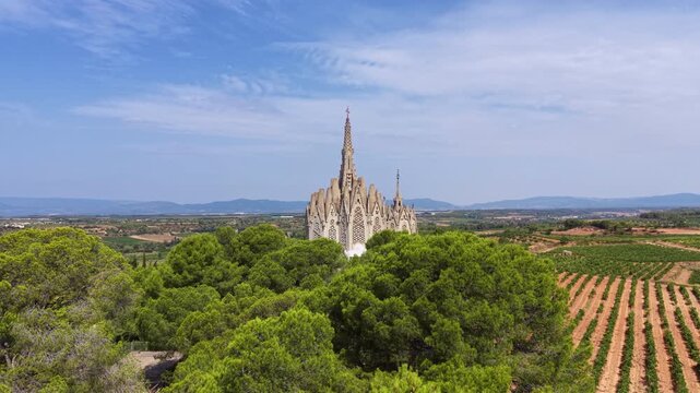Aerial view of Santuari de la Mare de Deu de Montferri rising above dense pine forest in Tarragona, Catalonia, its catenary arch spires surrounded by vineyards and farmland under a hazy blue sky