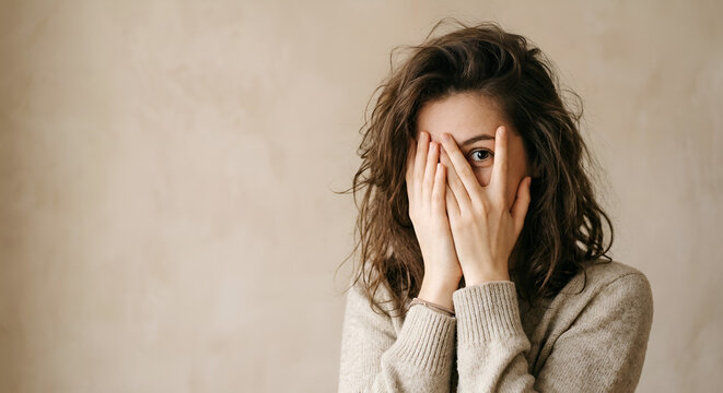 Young woman covering her face with hands and looking surprised indoors  