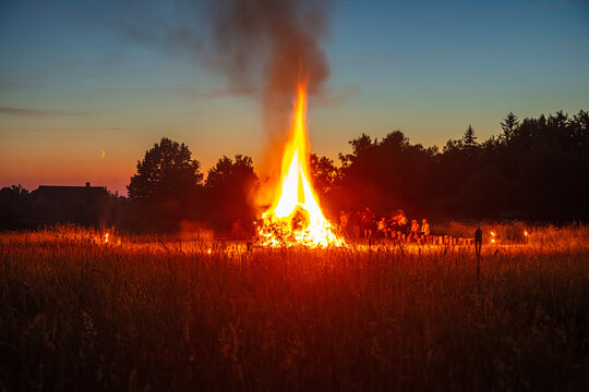 View of a large bonfire burning in a grassy field at dusk with silhouettes of people gathered around the flames and a crescent moon in the sky in Latvia.