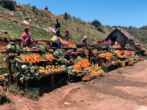 Madagascar - 19 November 2023: View of a vibrant roadside market with local vendors selling fresh produce like carrots, potatoes, and cabbage on wooden stalls against a hilly landscape.