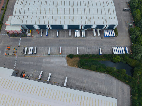 Aerial view of a large logistics warehouse facility with numerous semi-trailers parked at loading bays in a paved industrial yard near a small pond in Burton upon Trent, England, United Kingdom.
