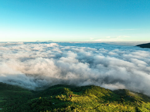 Aerial view of Gunung Mongkrang rising above a thick sea of clouds with lush green mountain slopes and clear blue sky in Jatiyoso, Central Java, Indonesia.