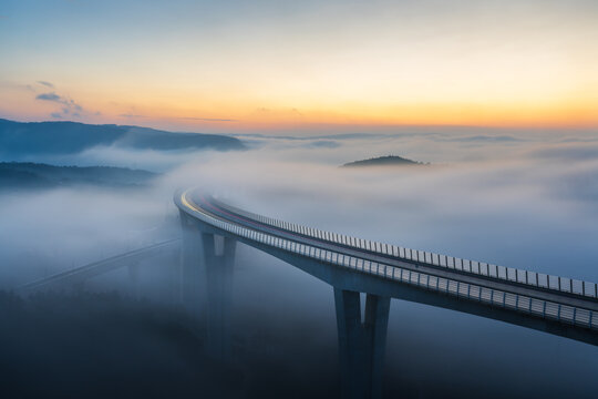 Aerial view of Crni Kal Viaduct emerging from thick morning fog under a golden sunrise sky in Crni Kal, Koper, Slovenia.