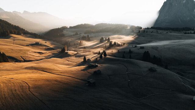 Aerial view of Alpe di Siusi with rolling alpine meadows, morning mist, and traditional wooden huts under golden sunlight in Bolzano, Trentino-Alto Adige, Italy.