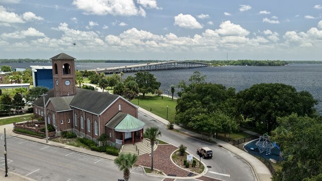 Aerial view of a historic brick church with a clock tower situated near the St. Johns River, featuring lush green trees and a bridge in the background in Palatka, Florida, United States.