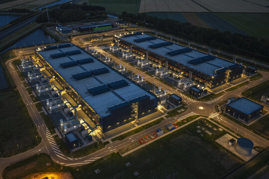 Aerial view of a large hyperscale data center illuminated at night, featuring expansive industrial buildings, cooling infrastructure, and surrounding road networks in the Netherlands.