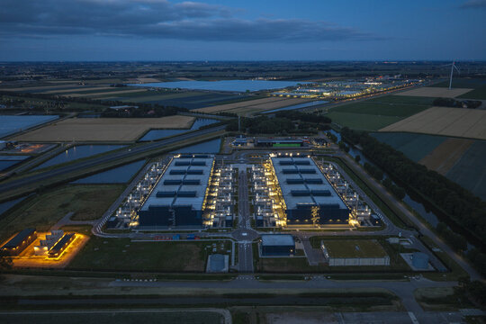Aerial view of a large hyperscale data center illuminated at dusk, surrounded by agricultural fields, canals, and roads in the Netherlands.