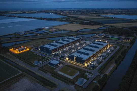 Aerial view of a hyperscale data center complex illuminated at dusk, surrounded by agricultural fields and waterways in the Netherlands.