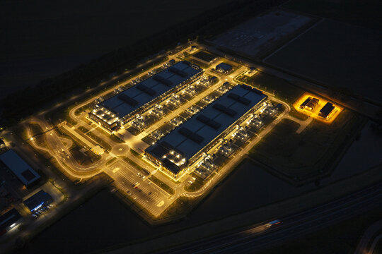 Aerial view of a large data center facility illuminated at night, featuring modern industrial buildings, parking areas, and surrounding roads in Middenmeer, North Holland, Netherlands.