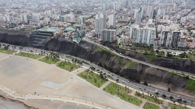 Aerial view of the Circuito de Playas highway running along the base of the cliffs with the urban skyline of high-rise buildings in the background in Magdalena del Mar, Provincia de Lima, Peru.