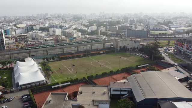 Aerial view of a sports complex with a soccer field, tennis courts, and a white tent structure, with a train passing on an elevated track in Santiago de Surco, Lima Province, Peru.