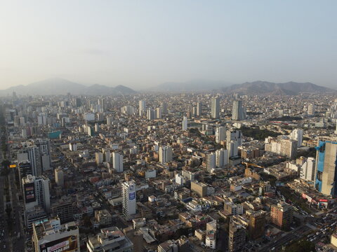 Aerial view of Bosque El Olivar surrounded by dense urban buildings, high-rise apartments, and distant mountains under a hazy sky in San Isidro, Provincia de Lima, Peru.