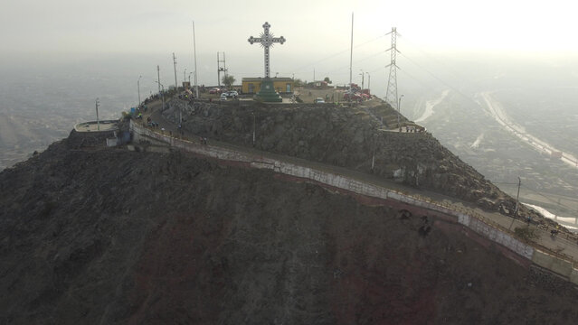 Aerial view of Cerro San Cristobal with its prominent cross, winding roads, and rocky terrain overlooking the sprawling urban landscape of Lima, Lima, Peru.