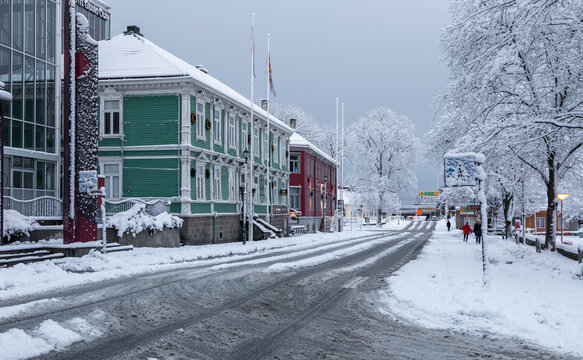 Trondheim, Norway - 30 December 2024: View of a snow-covered street in Trondheim featuring historic wooden buildings, winter trees, and tire tracks under an overcast sky.