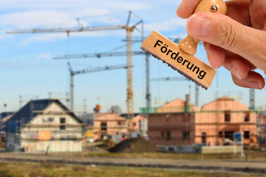 &Uuml;ber ein Neubaugebiet mit Wohnungen und H&auml;usern im Rohbau wird ein Holzstempel gehalten mit dem Aufdruck: F&ouml;rderung - FOTOMONTAGE