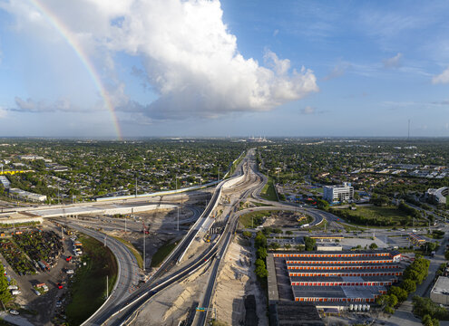 Aerial view of the Golden Glades Interchange under construction, featuring complex highway ramps, urban sprawl, and a rainbow arching across the sky. Miami, Florida, United States.