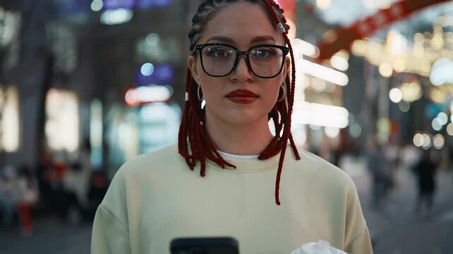 Young woman with red dreadlocks walks city street at night holding coffee and using phone