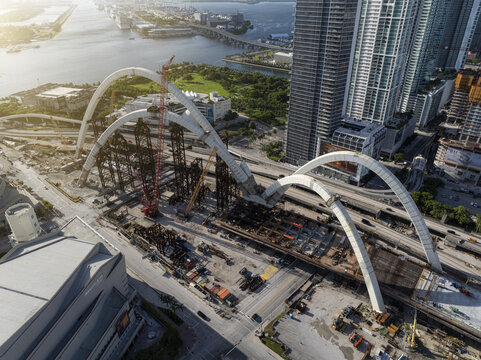 Aerial view of the Signature Bridge under construction featuring large white arches, construction cranes, and surrounding urban infrastructure in Miami, Florida, United States.