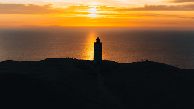 Aerial view of the iconic Rubjerg Knude lighthouse silhouetted against a vibrant golden sunset over the North Sea, with the surrounding sand dunes of Lokken, Denmark.