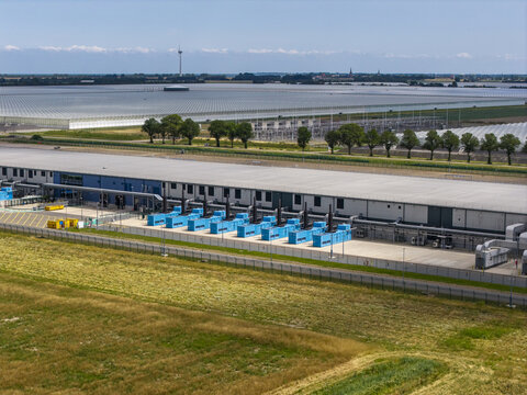 Aerial view of a modern data center facility with blue cooling units and a large industrial building situated among agricultural fields and greenhouses in the Netherlands.