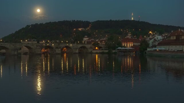 Charles Bridge and Vltava River in Prague at night under a bright full moon. City lights reflect on water near Petrin Hill. Scenic landscape for European travel, tourism and urban night life.