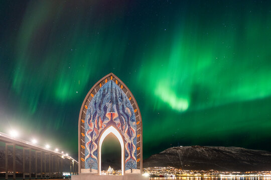 View of the vibrant green northern lights illuminating the night sky above the Tromso Bridge and a decorative arch monument in Tromso, Troms, Norway.