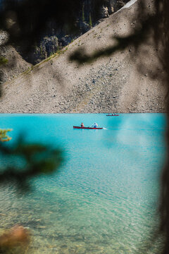 View of people paddling a red canoe on the turquoise glacial waters of Moraine Lake with rocky mountain slopes in the background in Improvement District No. 9, Alberta, Canada.
