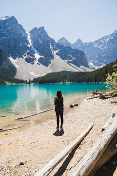 View of a woman with a backpack standing on the shore of Moraine Lake, with turquoise water and snow-capped mountains in the background in Improvement District No. 9, Alberta, Canada.