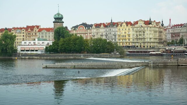 Water flows over a river weir on the Vltava River in Prague. Historic Sitkov Water Tower and colorful embankment buildings stand in the background. Ideal scene for European travel and tourism.