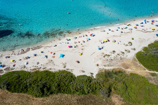 Aerial view of Plage de Ghjunchitu with turquoise water, white sand, and colorful beach umbrellas surrounded by lush green vegetation in Corbara, Corse, France.