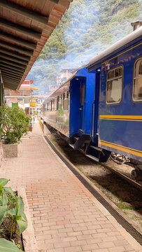 View of a blue passenger train parked at a brick platform with lush green mountains in the background in Cusco, Cusco, Peru.